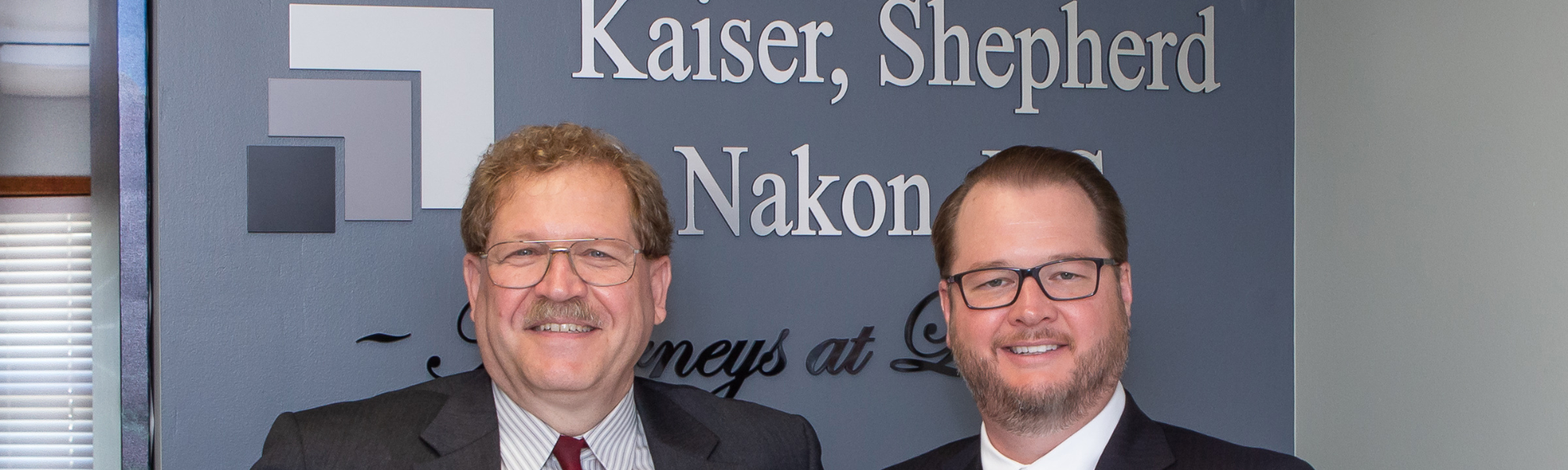 James W. Kaiser and Lehn Shepherd smiling in front of the Kaiser, Shepherd & Nakon, P.C. sign on a wall.